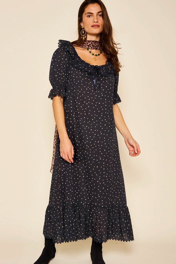 A woman stands against a plain background wearing the Aprilia - Starry Night Navy dress and accessorizes with earrings, a necklace, and a matching scarf from the Bobbie Bloom collection.