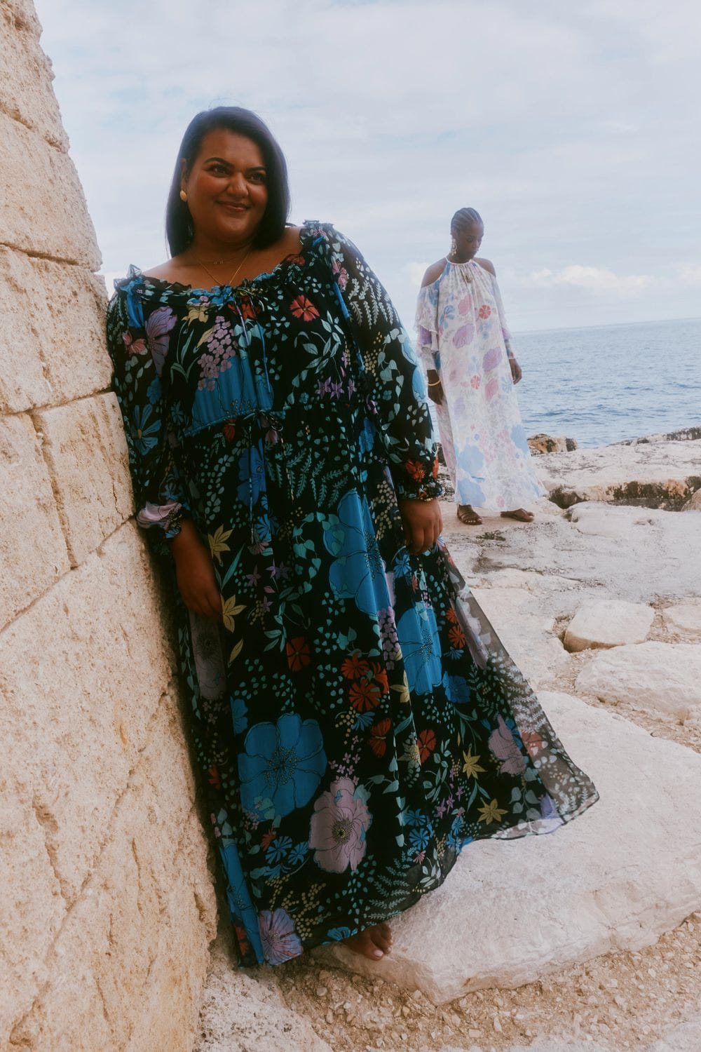 Woman in a floral dress standing by a stone wall with another person in the background.