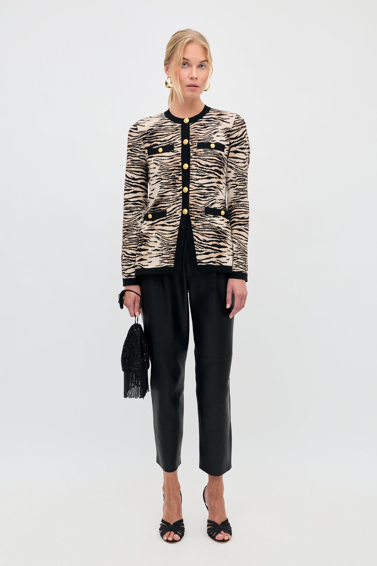 A model wearing the Raylene - Cooper Tiger Beige cardigan with black trim and gold buttons, black trousers, and heeled sandals, holding a small black beaded handbag.