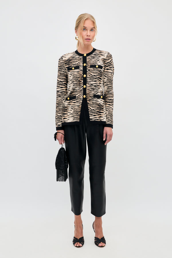 A model wearing the Raylene - Cooper Tiger Beige cardigan with black trim and gold buttons, black trousers, and heeled sandals, holding a small black beaded handbag.