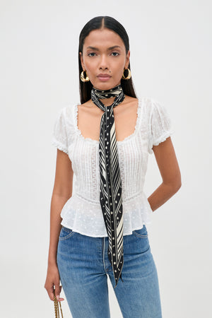 Woman wearing the Sandi - Ivory broderie anglaise cotton blouse with delicate lace inserts, blue jeans, gold hoop earrings, and a black-and-white patterned scarf tied around her neck, standing against a plain white background—effortlessly showcasing everyday style.