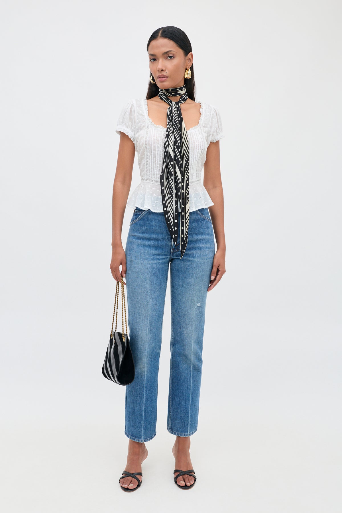 A woman stands against a white background wearing the Sandi - Ivory blouse with delicate lace inserts, paired with blue jeans, black sandals, a black and white patterned scarf, gold earrings, and holding a black purse—an effortlessly chic look perfect for everyday style.
