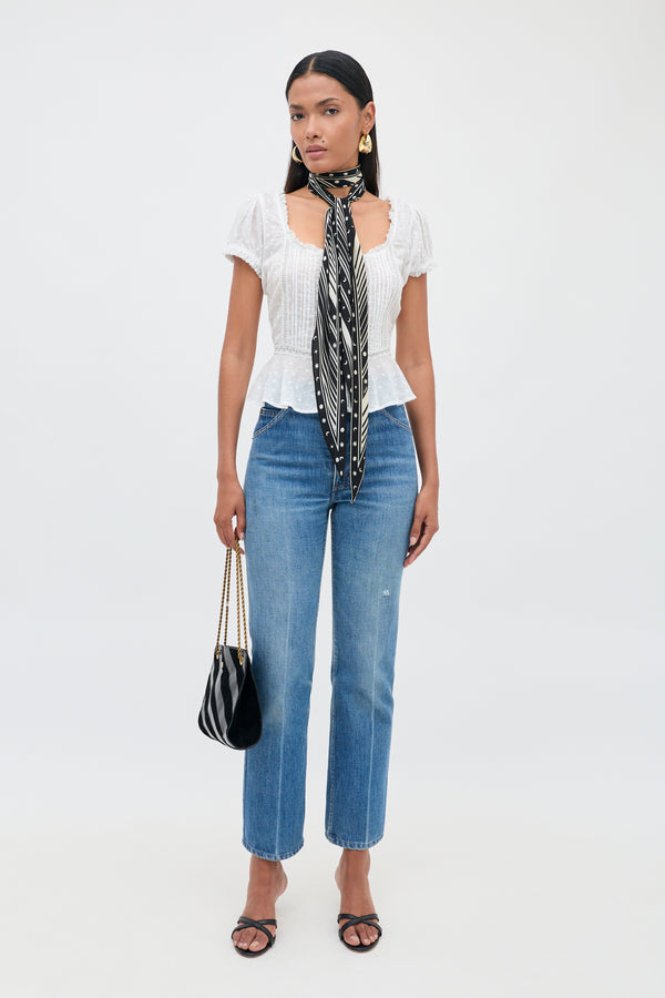 A woman stands against a white background wearing the Sandi - Ivory blouse with delicate lace inserts, paired with blue jeans, black sandals, a black and white patterned scarf, gold earrings, and holding a black purse—an effortlessly chic look perfect for everyday style.