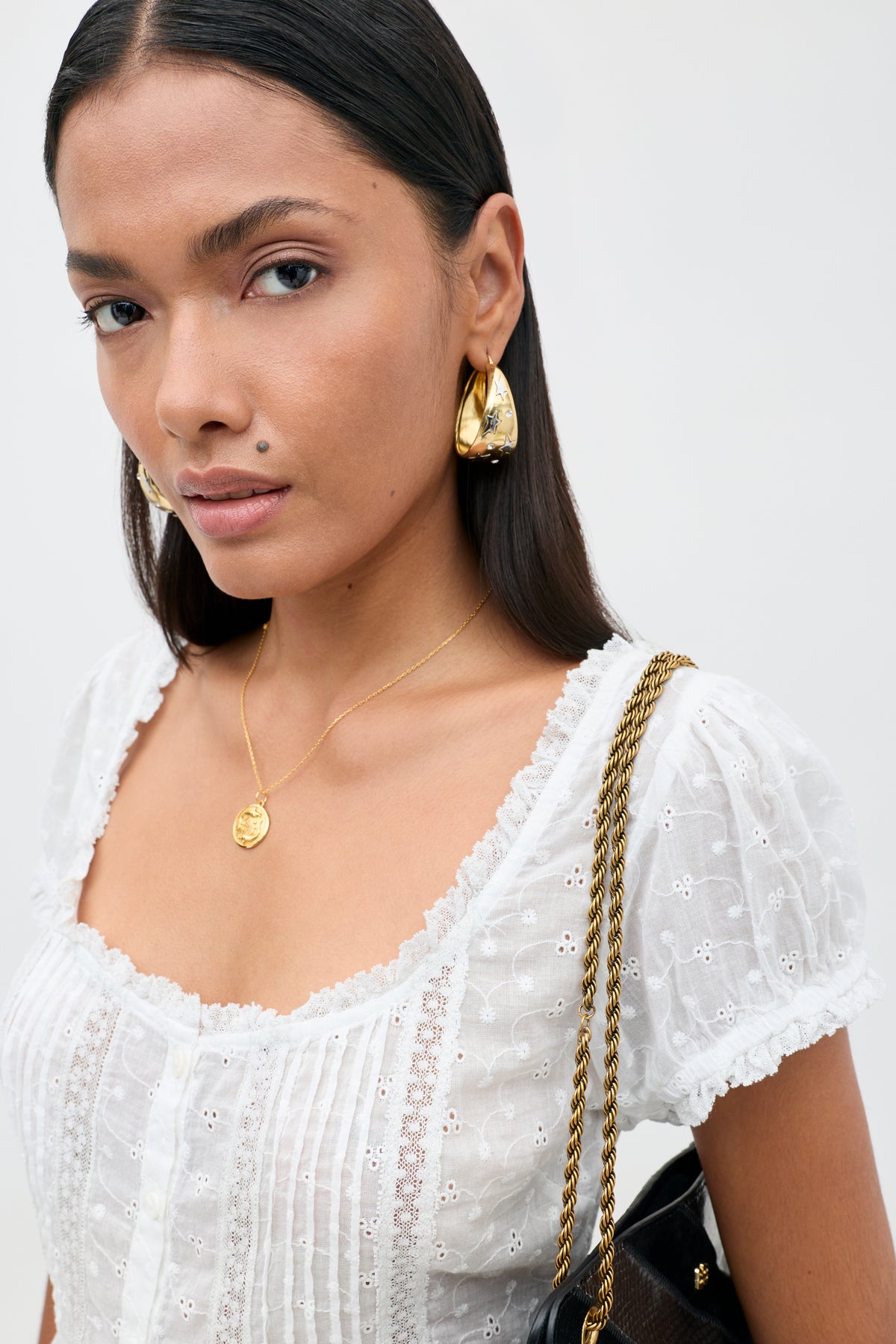 A woman with long dark hair wears gold earrings, a gold pendant necklace, and a white textured blouse with delicate broderie anglaise cotton and lace trim inserts. She carries the Sandi - Ivory bag with its distinctive gold chain strap over her shoulder and looks confidently at the camera, embodying effortless everyday style.