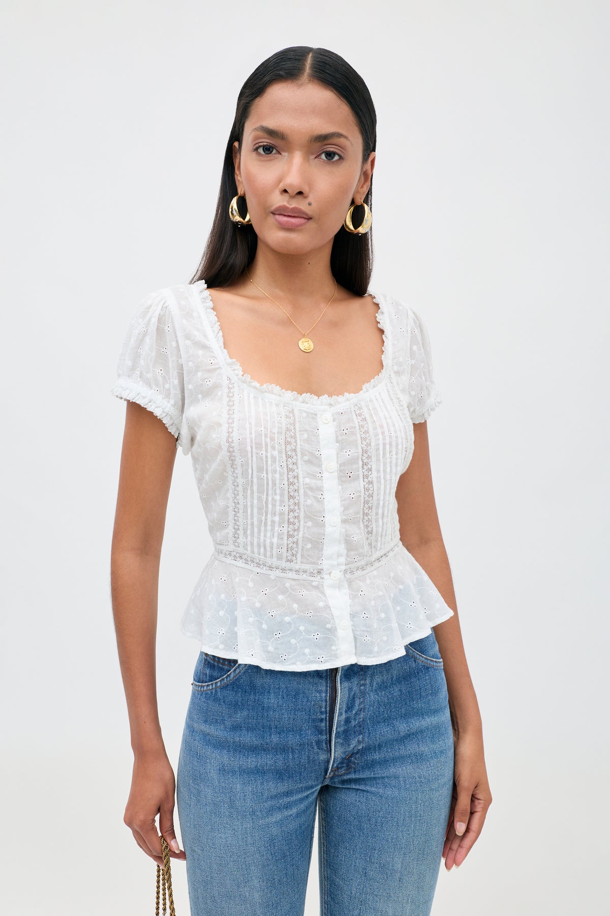 A woman with straight dark hair wears the Sandi - Ivory blouse—a white, short-sleeve, embroidered top crafted from broderie anglaise cotton with a peplum hem—paired with blue jeans, gold hoop earrings, and a gold pendant necklace, embodying effortless everyday style as she stands against a plain light background.