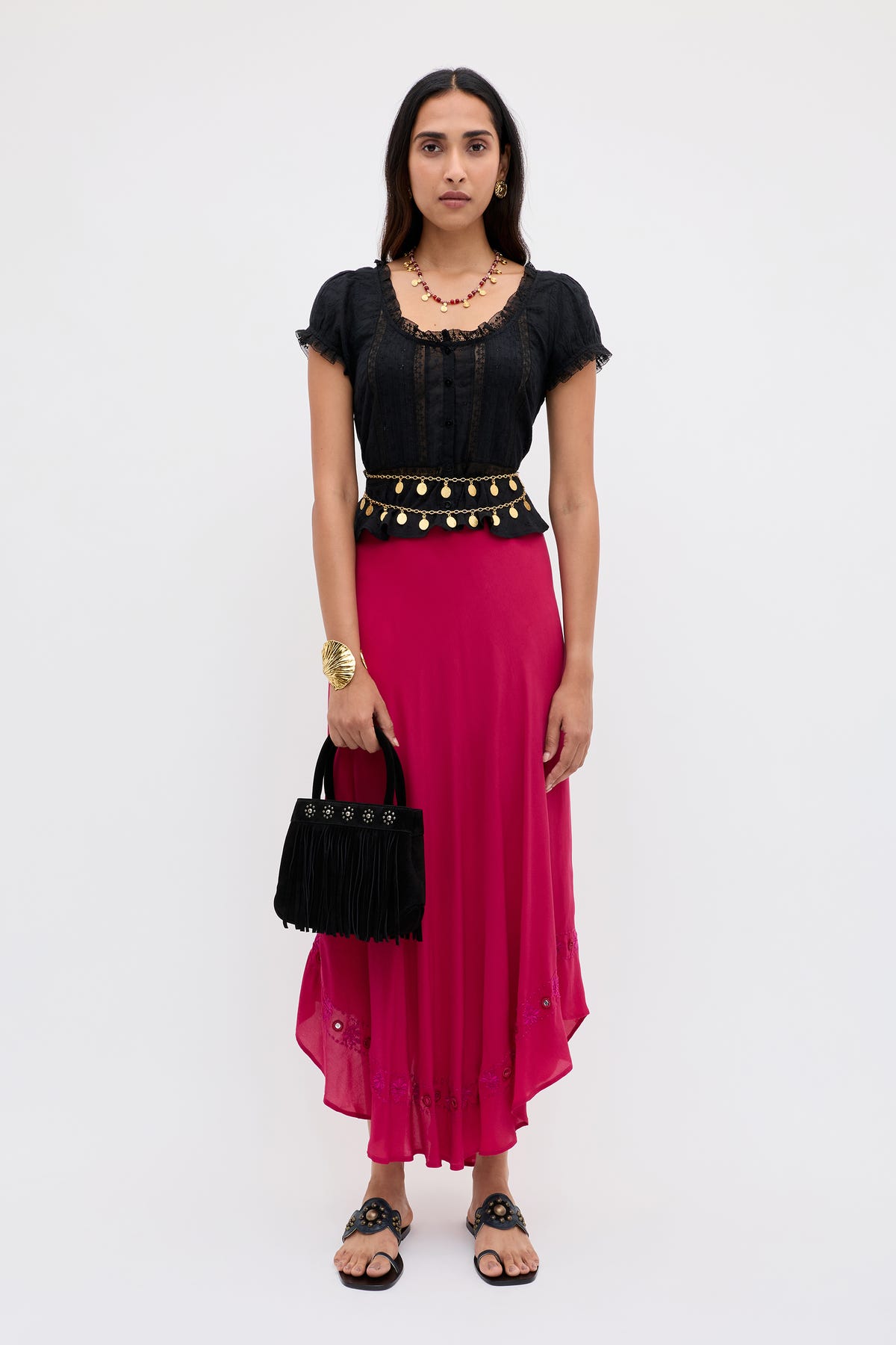 A woman stands facing forward against a plain light background, wearing the Rudy - Mirror Embellishment Magenta Pink skirt. She pairs it with a black top, black sandals, jewelry, and holds a black fringed handbag.