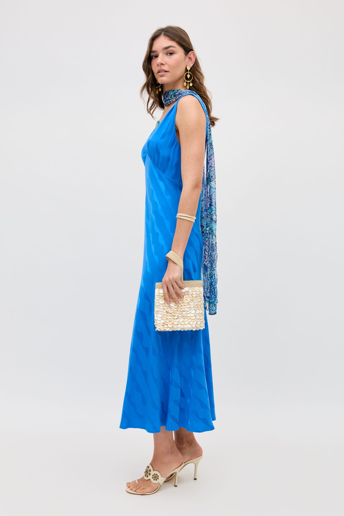 A woman in the Sandrine - Wave Jacquard Blue dress by RIXO, a standout transitional piece, styled with a patterned scarf draped over one shoulder, gold earrings and bracelet, heeled sandals, and a textured cream clutch.