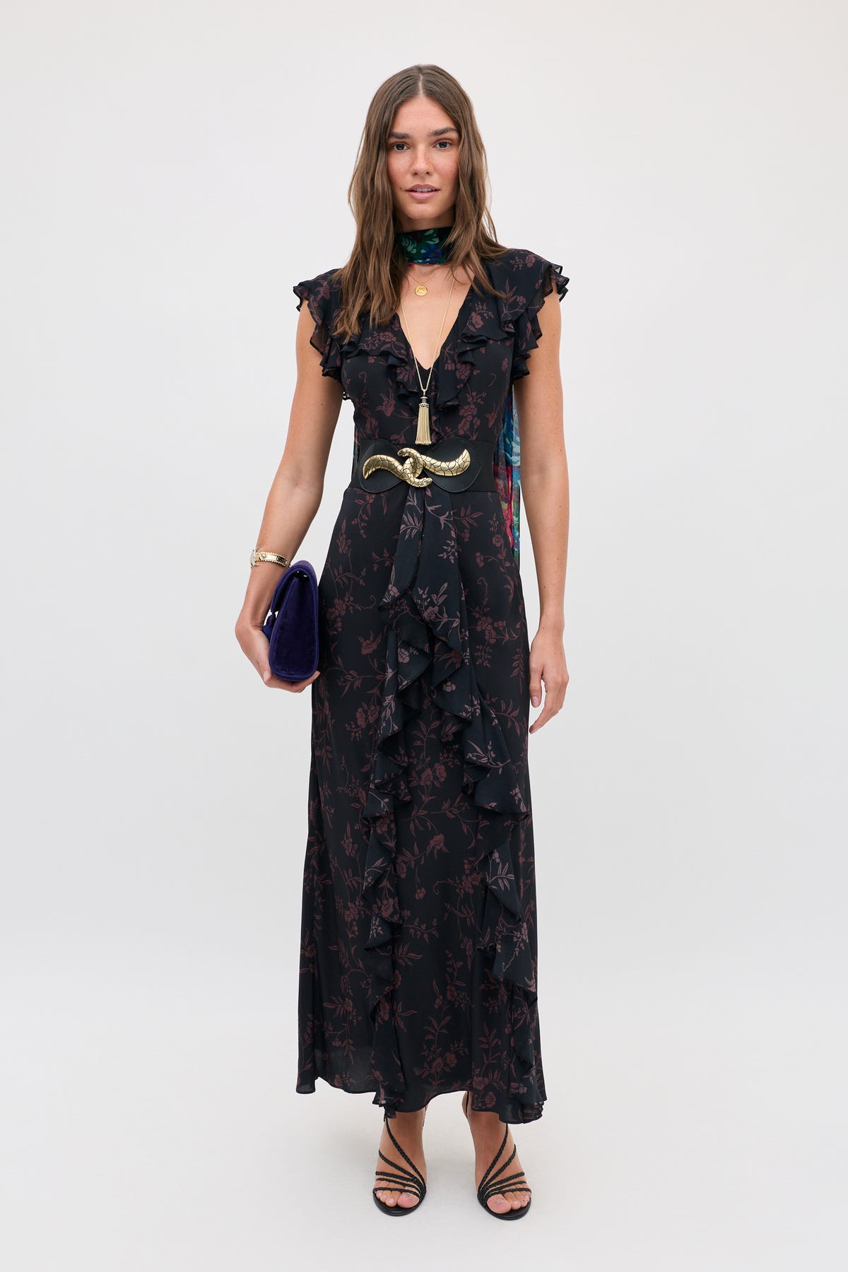 A model wears the Sofiya - Botanical Red, a long, sleeveless black dress with a red floral pattern, ruffled edges, and a gold-buckled waist belt, while holding a dark blue clutch.