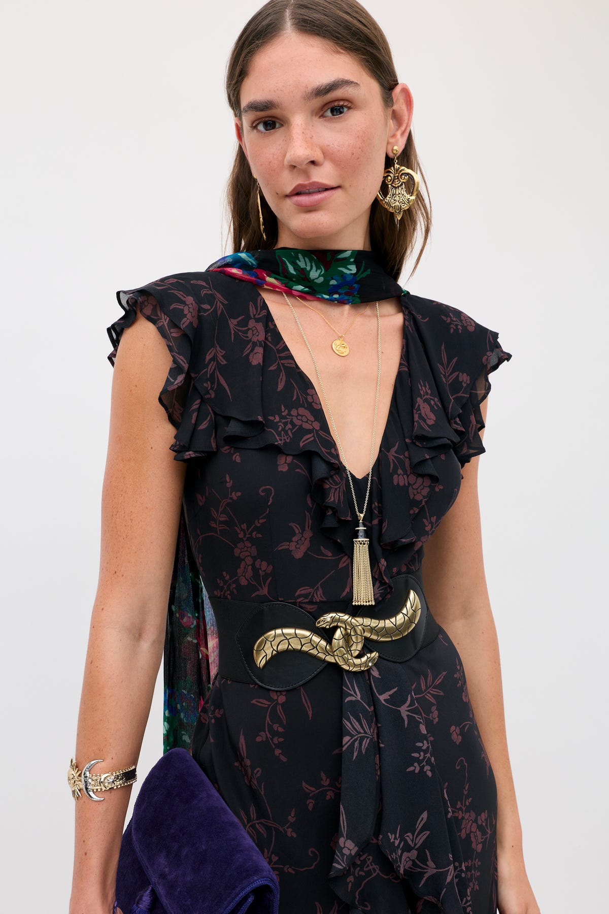 A person wears the Sofiya - Botanical Red, a black V-neck dress with a dark floral pattern, a wide black belt with a gold snake buckle, a colorful patterned scarf, and gold jewelry.