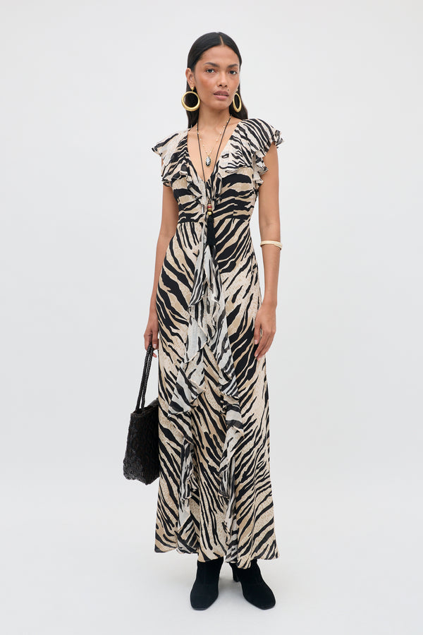 A woman stands against a plain background wearing the Sofiya - Cooper Tiger Beige long zebra-print dress, paired with black boots, large hoop earrings, and holding a black textured handbag. She has straight hair and a neutral expression.