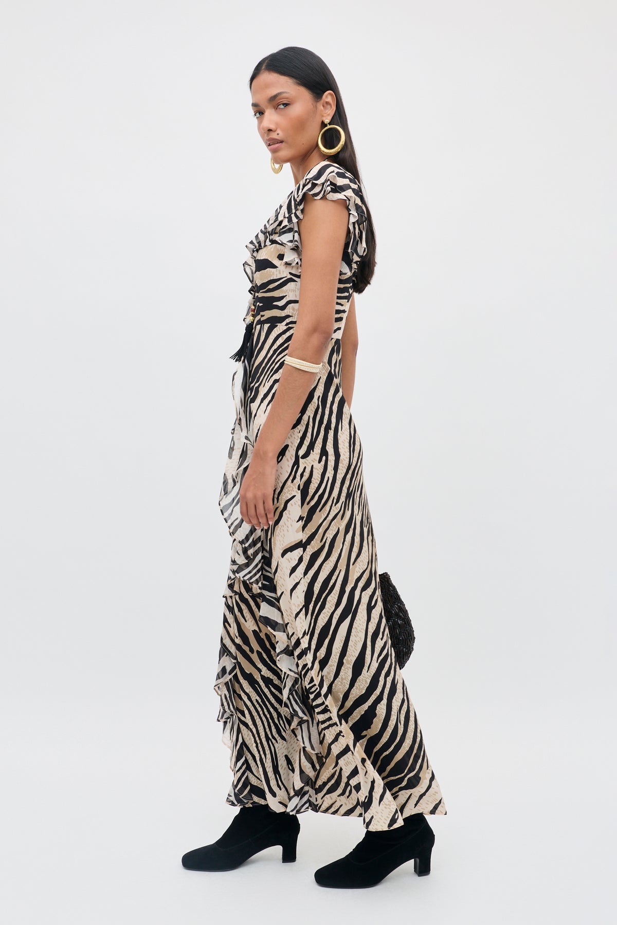 A woman wearing the Sofiya - Cooper Tiger Beige dress, paired with black ankle boots, large gold hoop earrings, a black bracelet, and holding a textured black bag, stands against a plain white background.