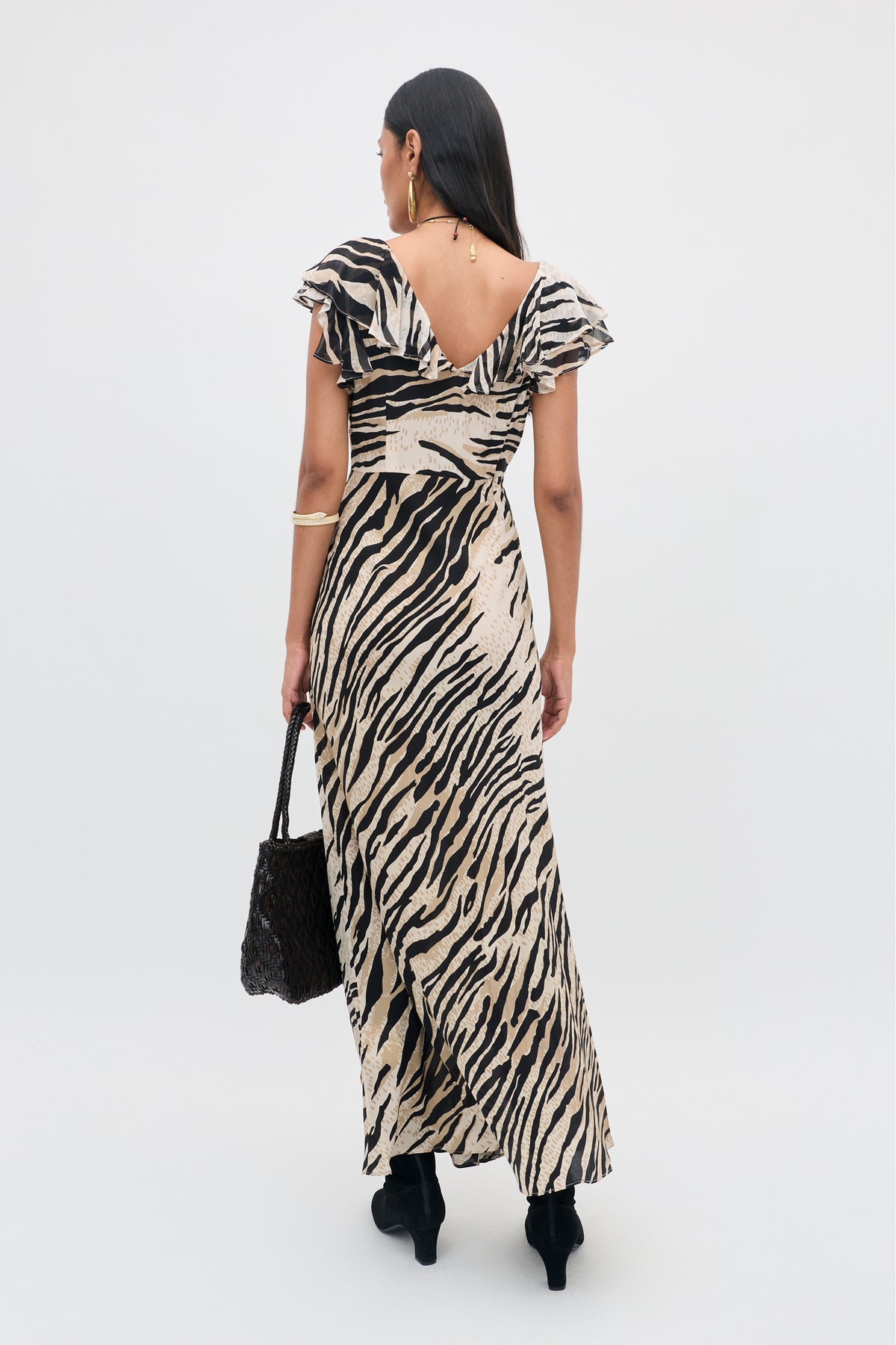 A woman with long dark hair wears the Sofiya - Cooper Tiger Beige maxi dress and black boots, holding a black textured handbag. She is standing with her back to the camera against a plain light background.