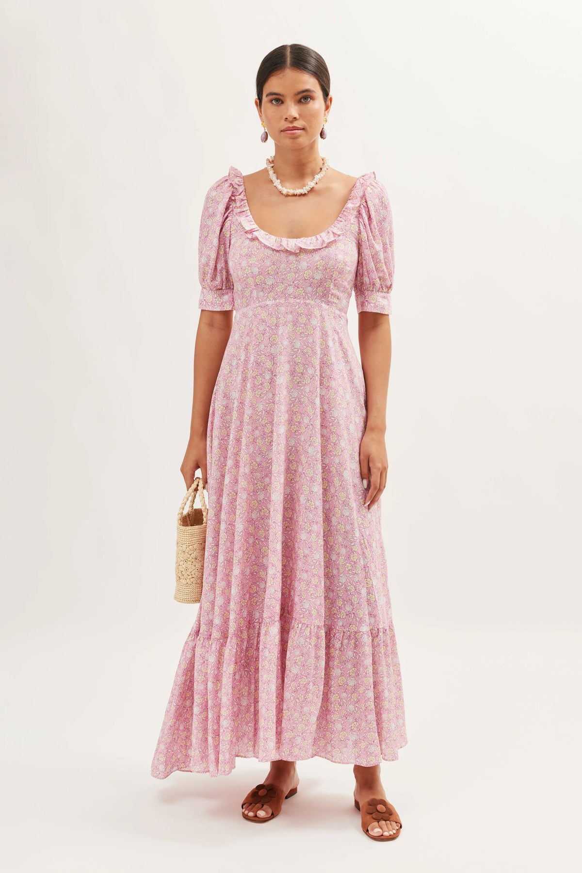 A woman stands against a plain background wearing the Sorana - Shell Pansy Blush dress with waist ties, puff sleeves, and a ruffled neckline. She holds a woven handbag, wears brown sandals, and a chunky necklace.