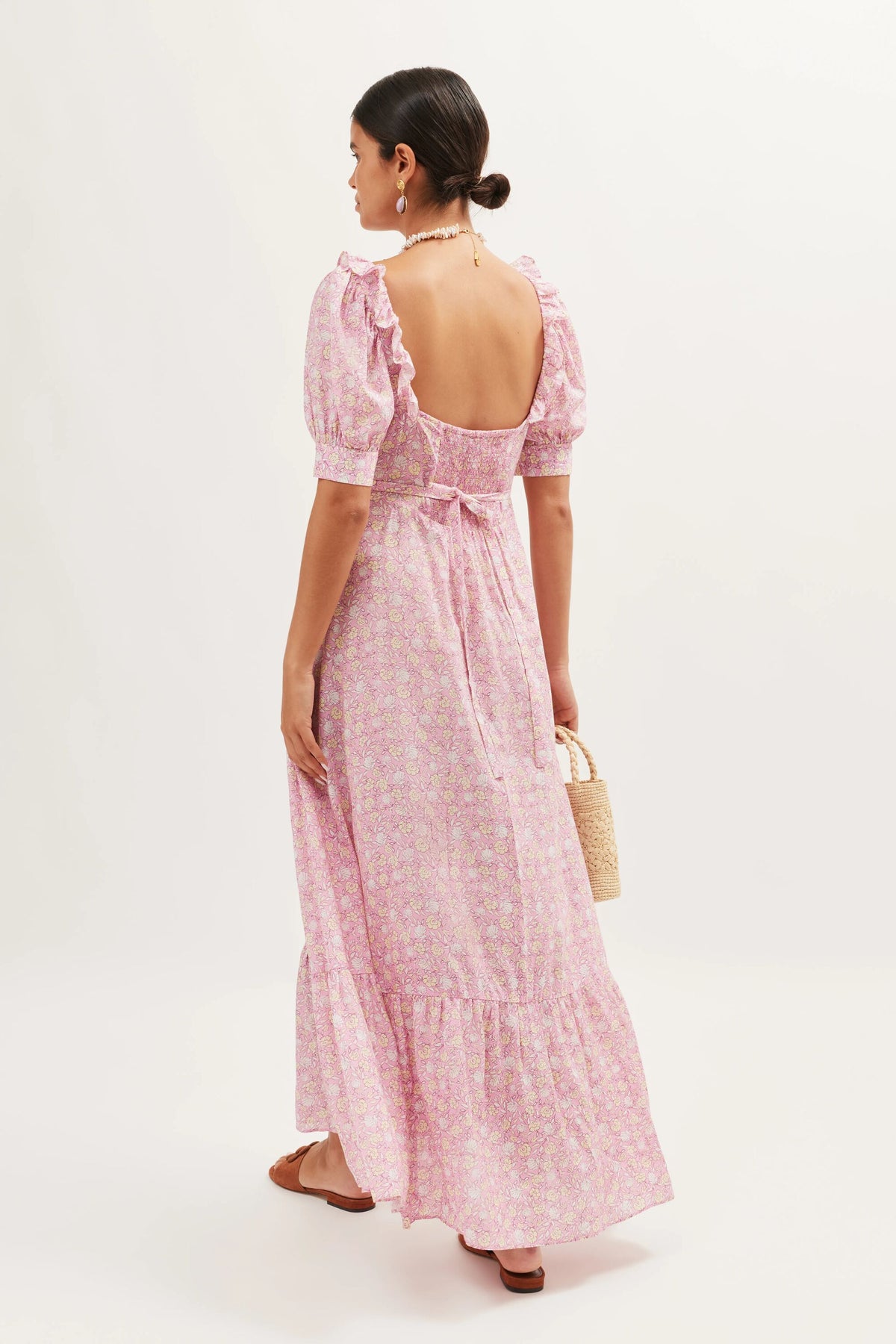 A woman faces away in the Sorana - Shell Pansy Blush, a light pink floral vintage prairie dress with puff sleeves, waist ties, and an open back. She holds a small woven handbag and wears flat sandals; her hair is in a low bun.