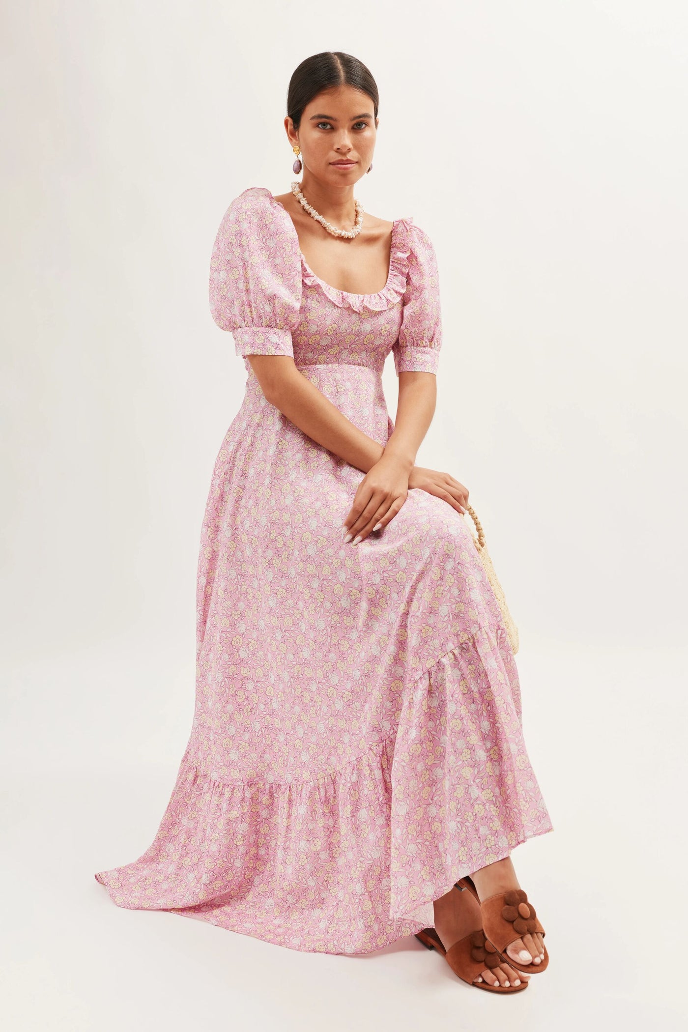 A woman sits on a stool wearing the Sorana - Shell Pansy Blush dress, a pink floral vintage prairie style with puffed sleeves, ruffled details, and a square neckline. She pairs it with pearl jewelry and brown sandals against a plain light background.