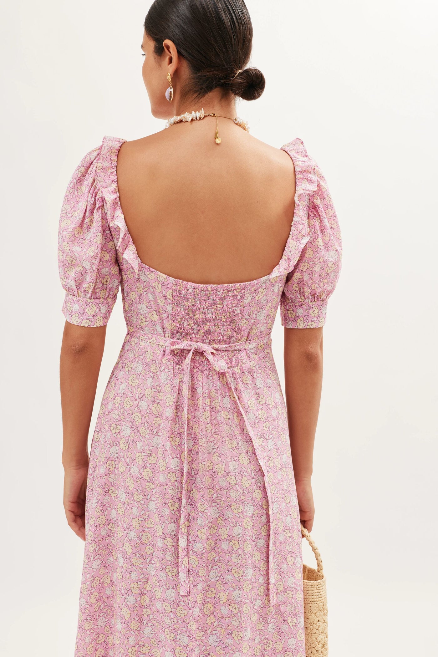 Wearing the Sorana - Shell Pansy Blush dress with puff sleeves, ruffled details, waist ties, and open back, she styles her hair in a low bun, adds earrings and a necklace, and carries a woven straw bag.