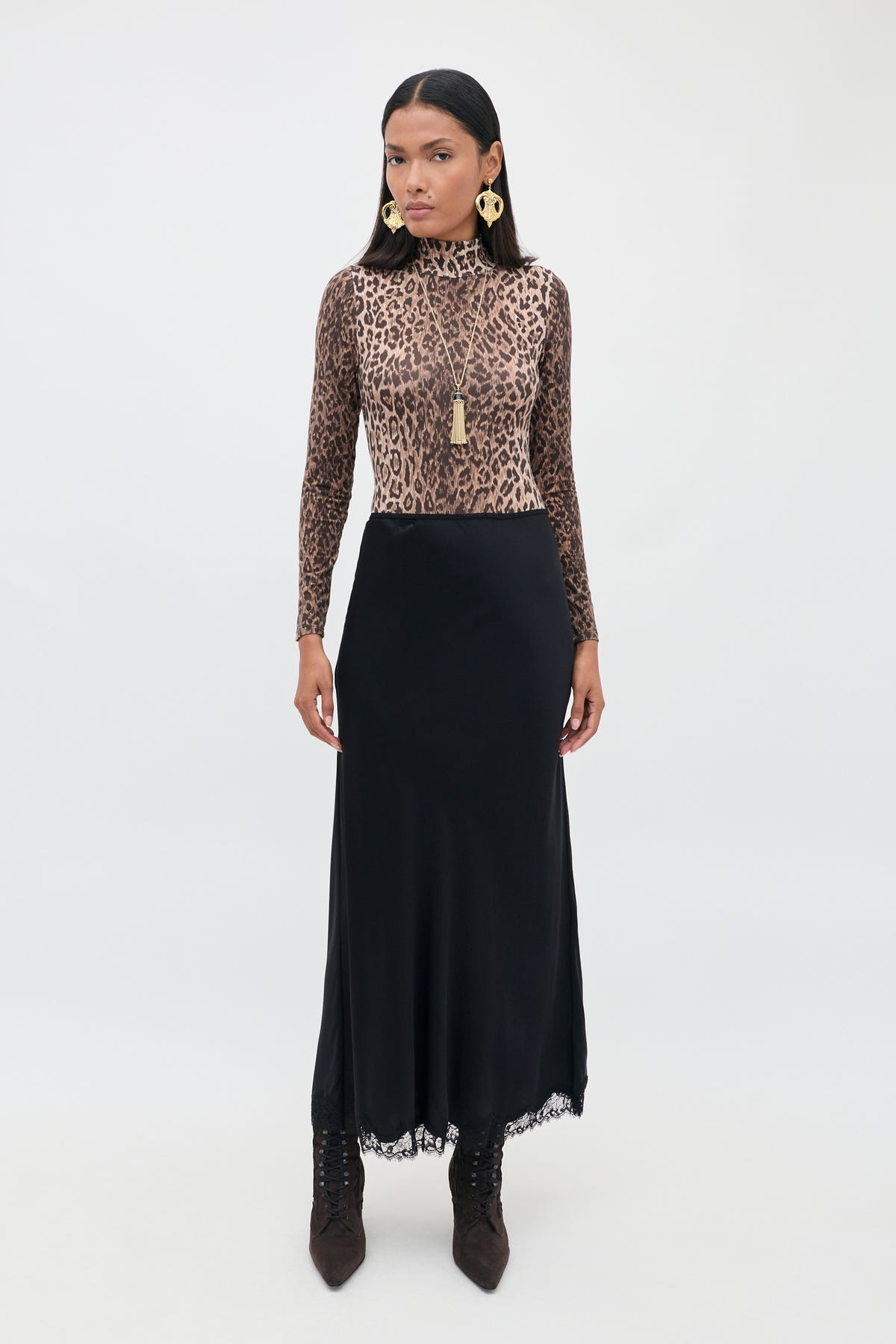 A woman stands against a plain background, wearing the Stassie - Black: a long matte satin skirt with delicate wavy lace trim, paired with a long-sleeve leopard print top, brown boots, gold earrings, and a necklace. She faces forward with a neutral expression.