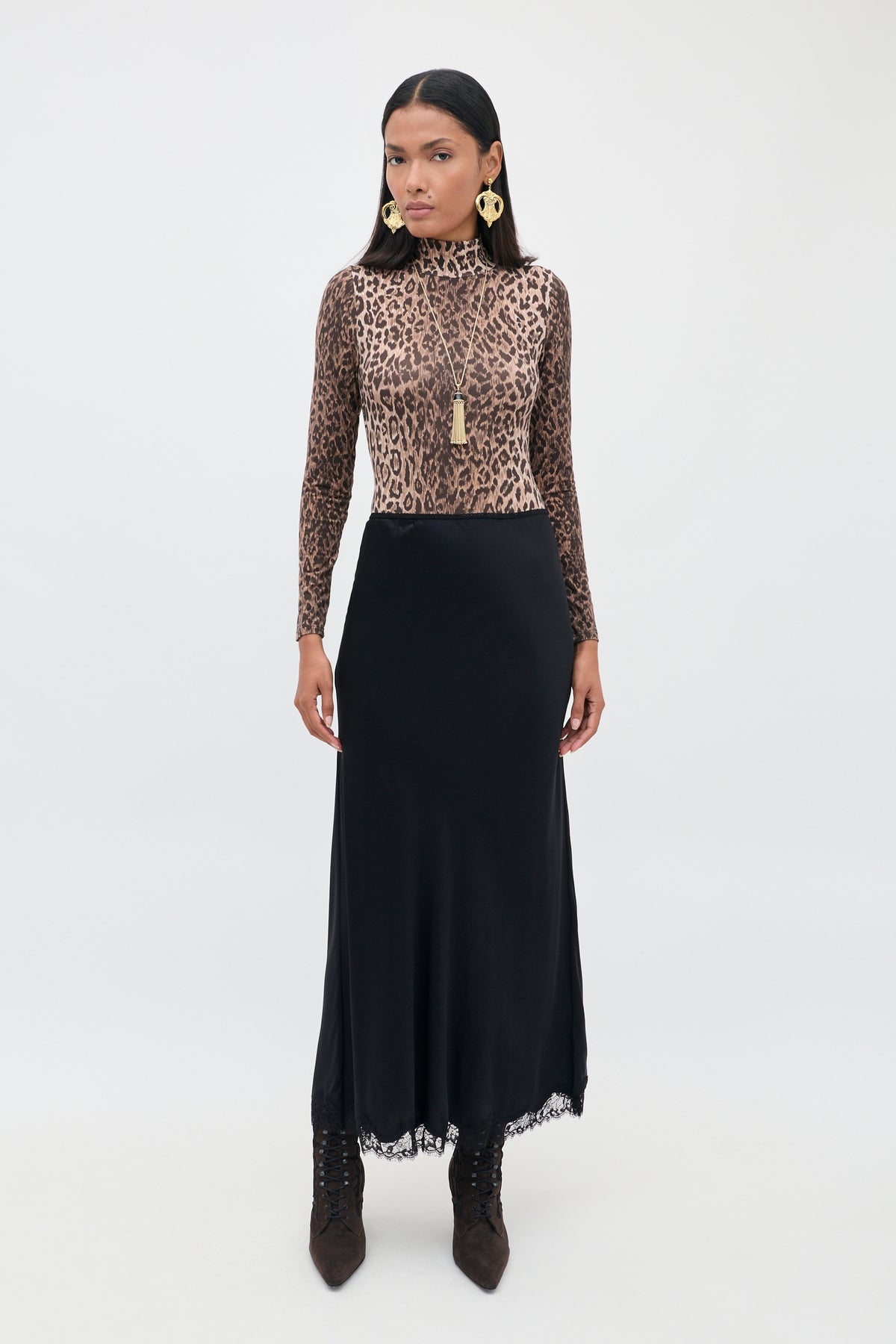 A woman stands against a plain background, wearing the Stassie - Black: a long matte satin skirt with delicate wavy lace trim, paired with a long-sleeve leopard print top, brown boots, gold earrings, and a necklace. She faces forward with a neutral expression.