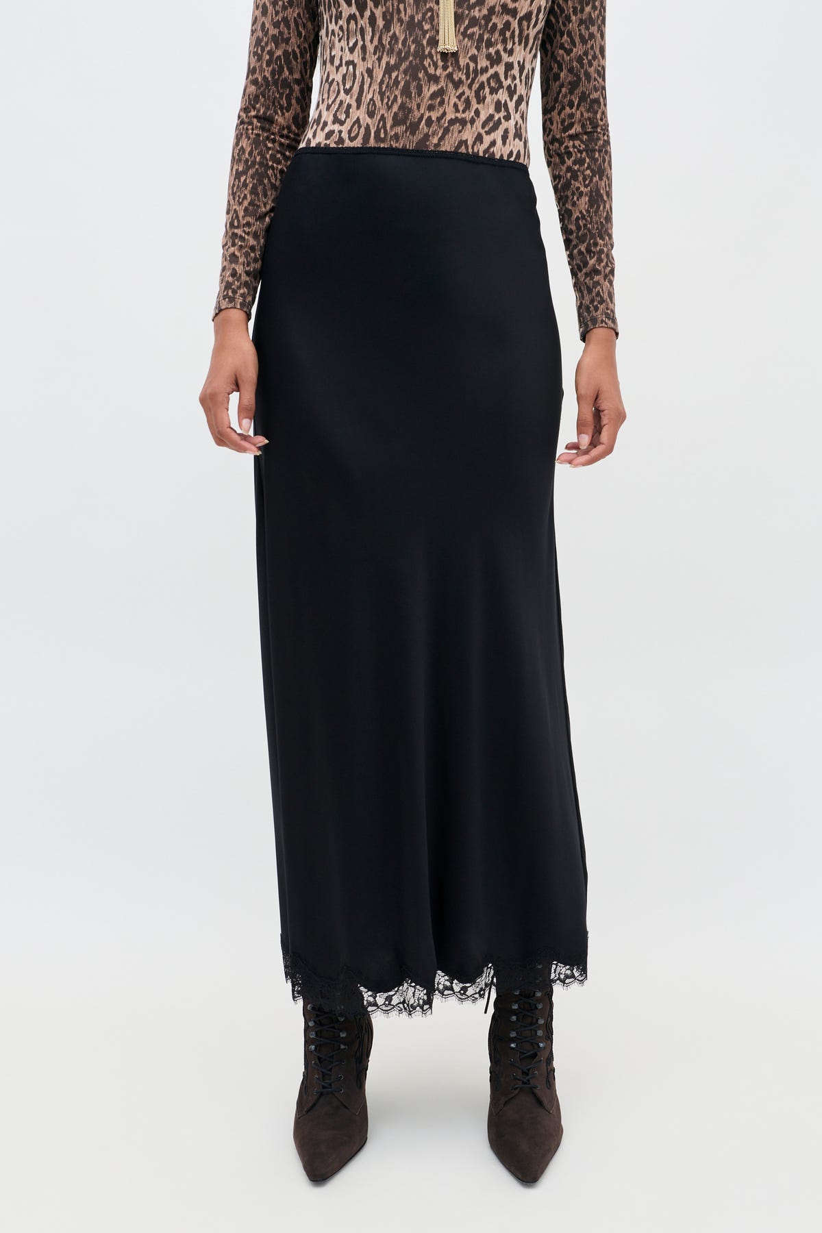 A person wearing the Stassie - Black long matte satin skirt with delicate wavy lace trim, a leopard print long-sleeve top, and pointed brown lace-up boots, standing against a plain background.