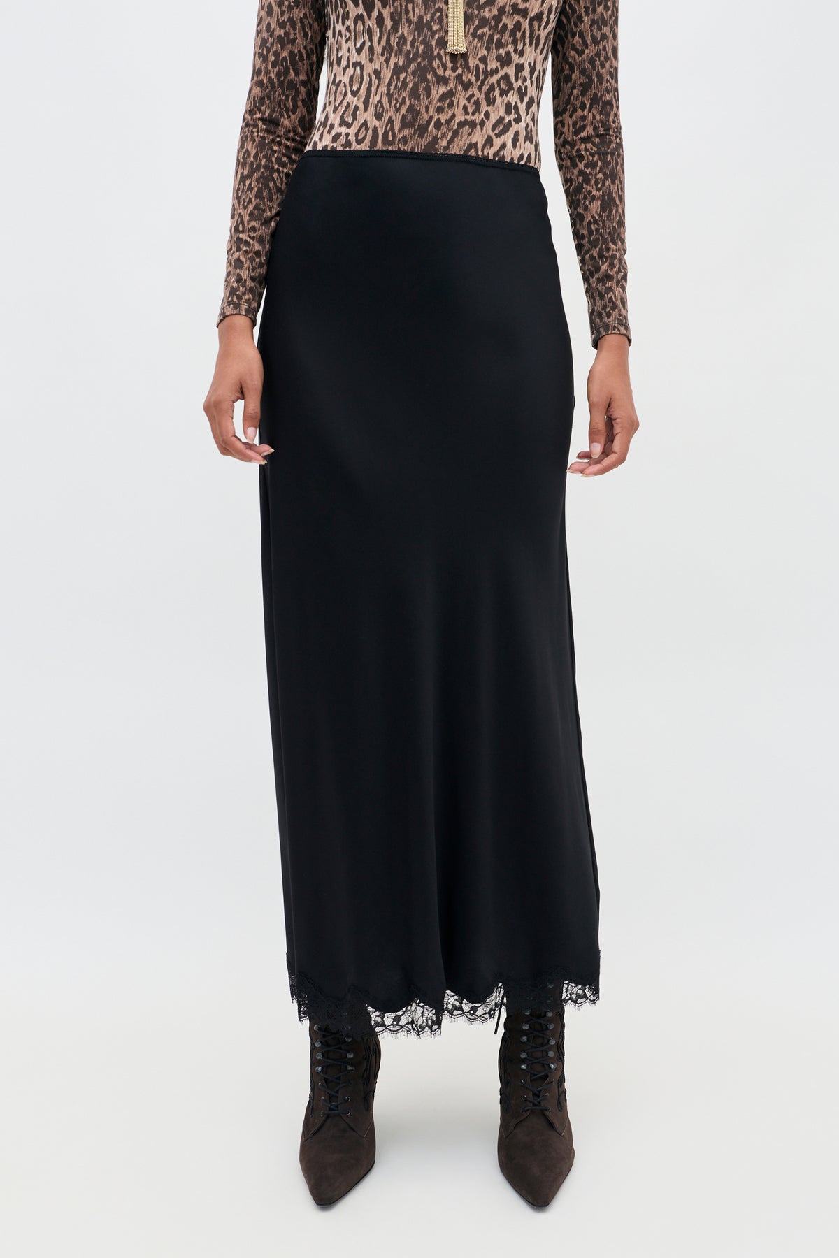 A person wearing the Stassie - Black long matte satin skirt with delicate wavy lace trim, a leopard print long-sleeve top, and pointed brown lace-up boots, standing against a plain background.
