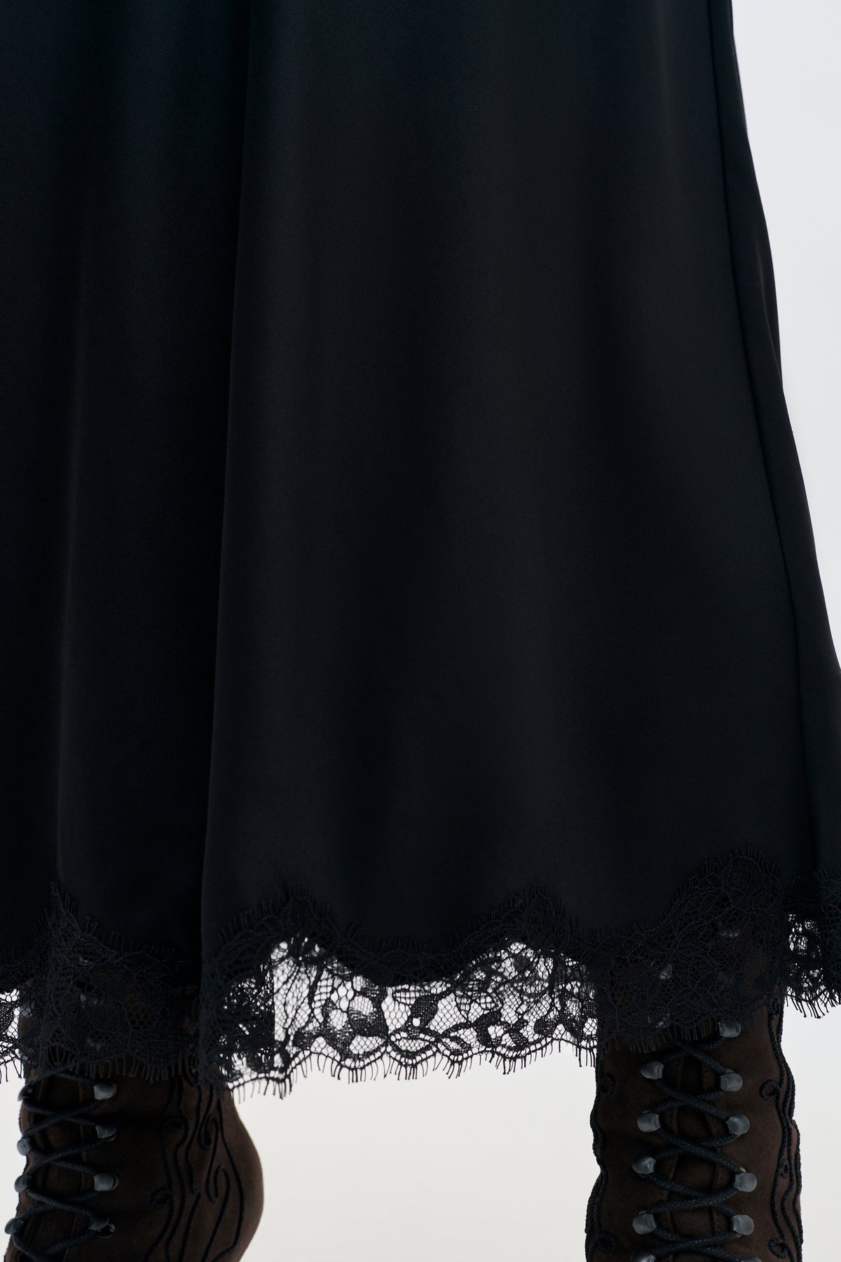 A close-up of the lower half of the Stassie - Black matte satin skirt with Paris lace trim at the hem, paired with dark lace-up boots.