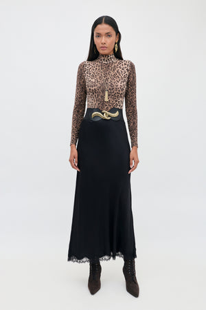 A woman stands facing forward against a plain white background, wearing the Stassie - Black matte satin skirt with wavy lace trim, paired with a long-sleeve leopard print top, a black belt featuring a gold snake buckle, brown boots, and gold earrings.