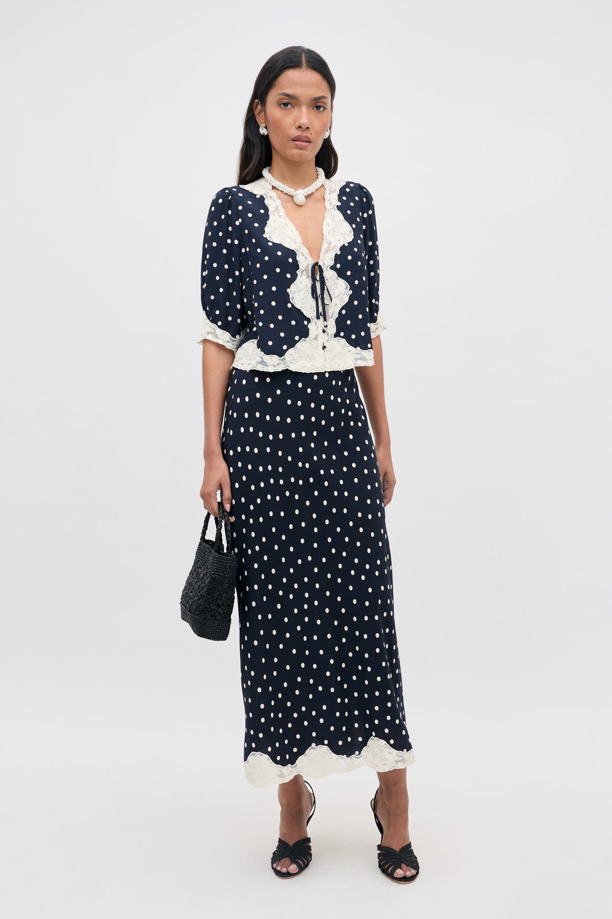 A woman with long dark hair and a neutral expression stands against a plain background wearing the Stassie - Layla Dot Navy two-piece outfit, which features navy blue polka dots and white lace trim. She completes her look with black sandals and a matching black handbag.