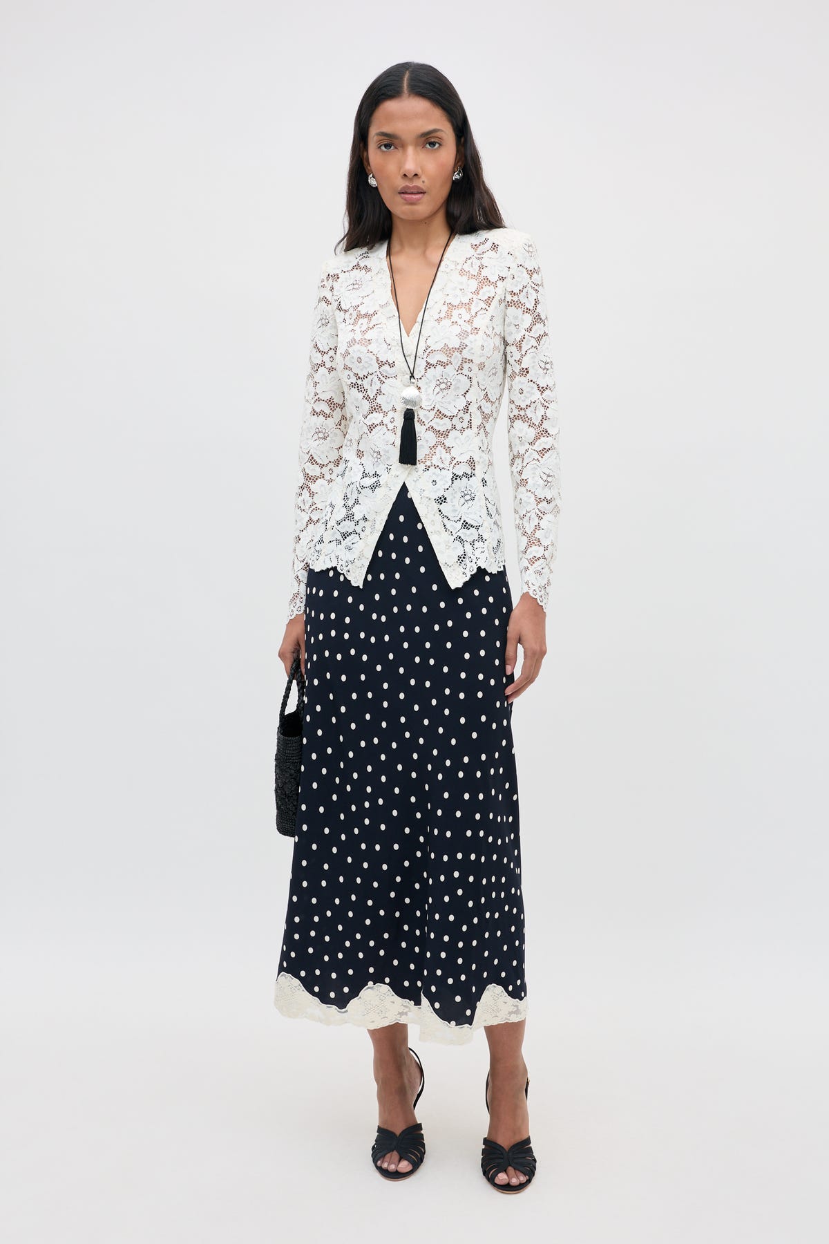 A woman stands against a plain background, wearing the Stassie - Layla Dot Navy midi skirt with black and white polka dots, paired with a white lace blazer, black sandals, and a long necklace. She holds a black handbag and has a neutral expression.