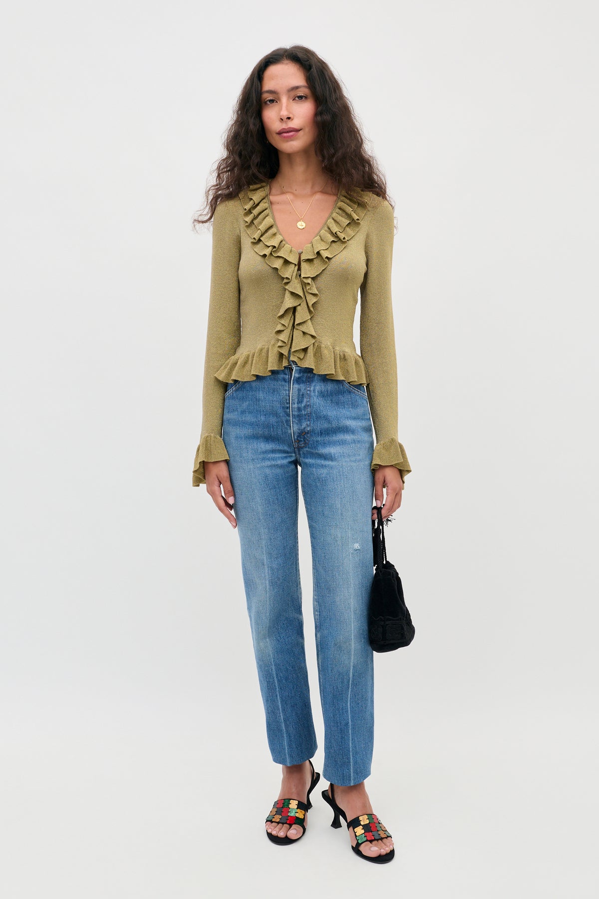 A person wearing a ruffled olive green long-sleeve top, blue straight-leg jeans, and Tacita - Gold patterned slides, holding a small black handbag.