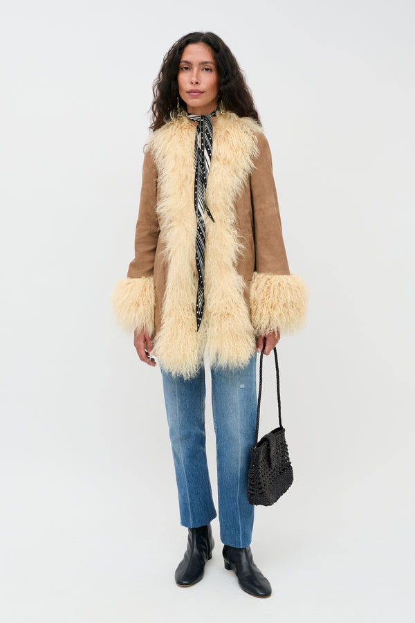 Wearing the Nellie - Tan suede coat with fluffy cream trim, blue jeans, black ankle boots, a black patterned scarf, and carrying a black woven bag, a person stands against a plain white background.