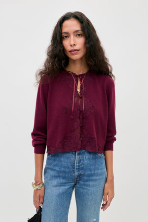 A woman with long curly hair is wearing the Tassia - Burgundy sweater, featuring lace details, paired with blue jeans, layered necklaces, and bracelets while standing against a plain light background.