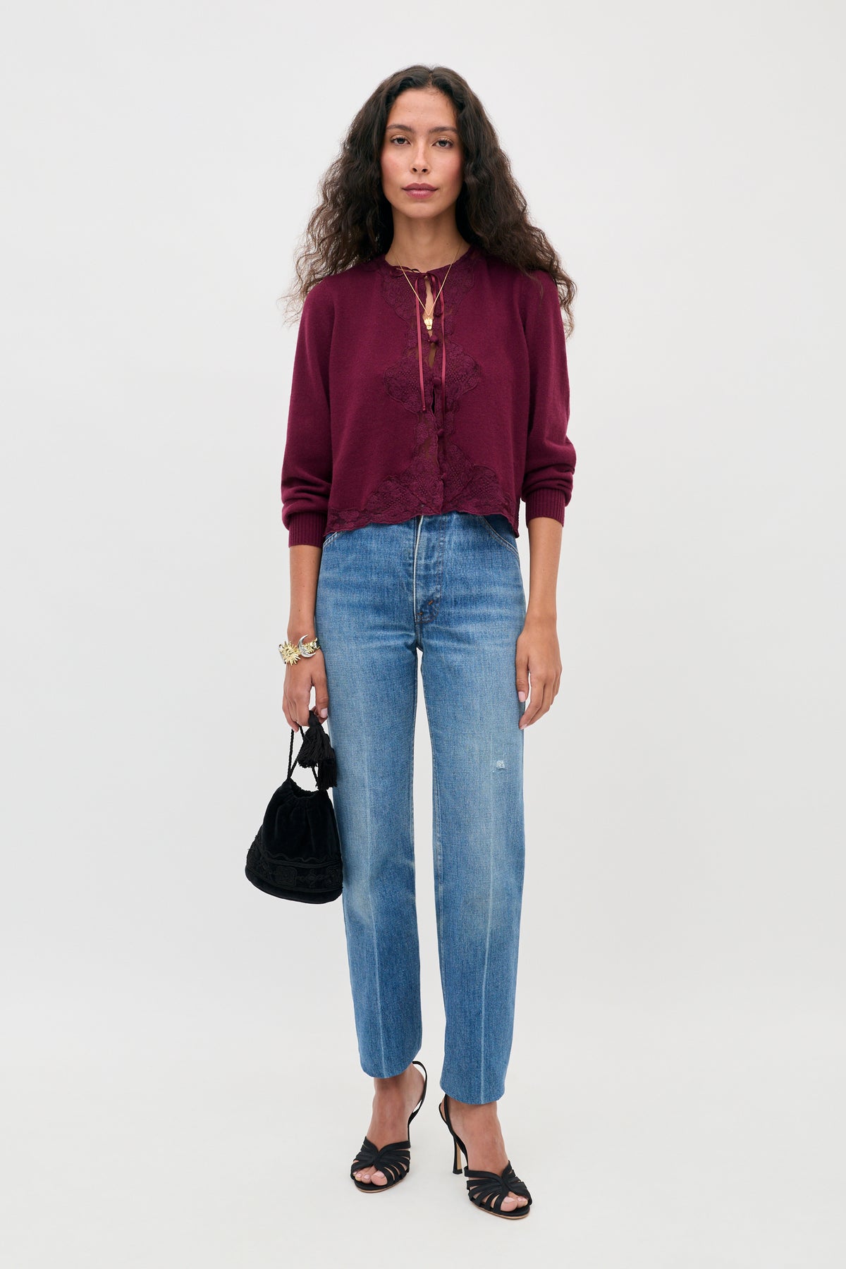 A woman stands against a plain background wearing the Tassia - Burgundy cardigan, blue jeans, black heeled sandals, and holding a small black handbag. She has long, curly hair and wears jewelry.