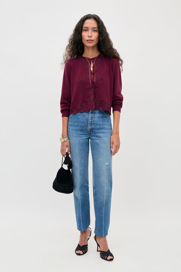 A woman stands against a plain background wearing the Tassia - Burgundy cardigan, blue jeans, black heeled sandals, and holding a small black handbag. She has long, curly hair and wears jewelry.
