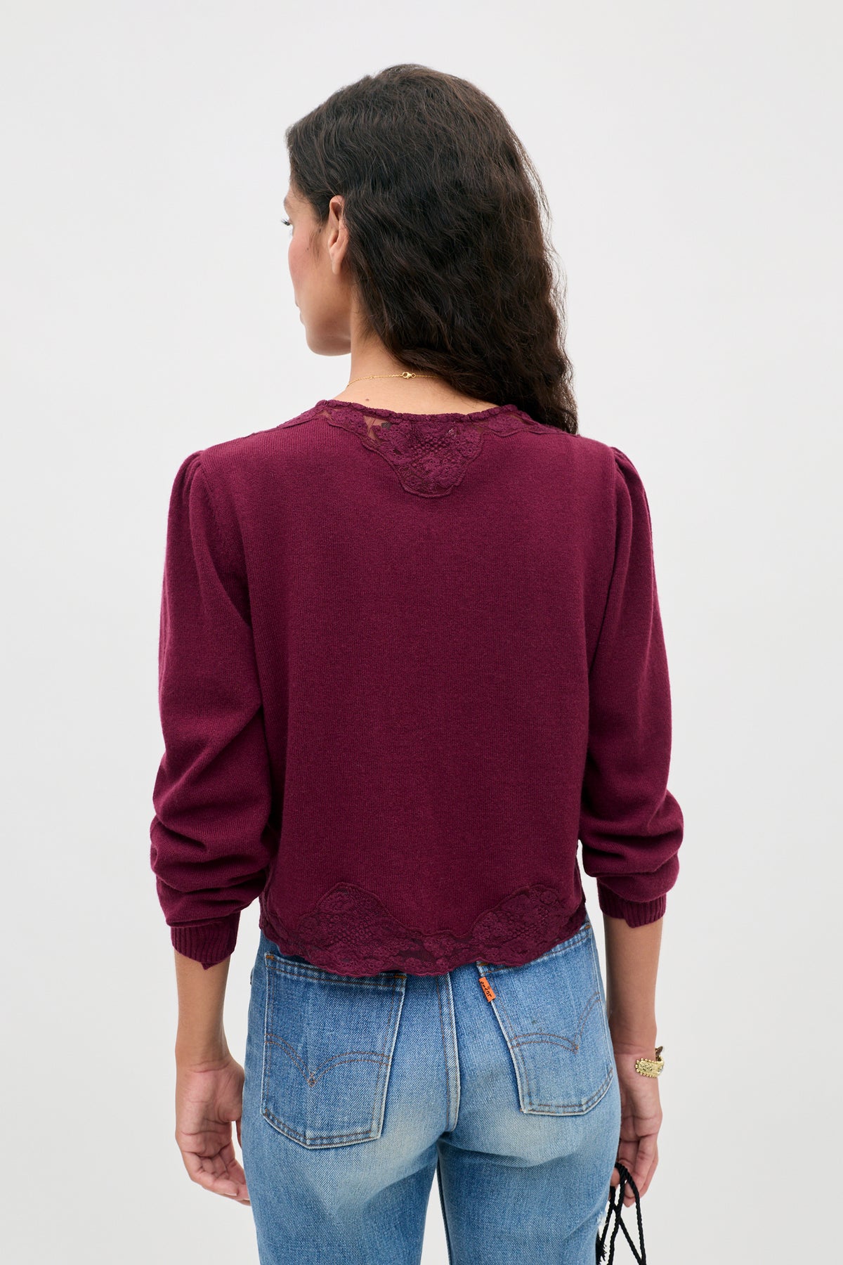 A person with long, wavy brown hair is wearing the Tassia - Burgundy sweater, featuring lace details, paired with blue jeans. The individual is facing away, showcasing the back of the outfit against a plain light background.