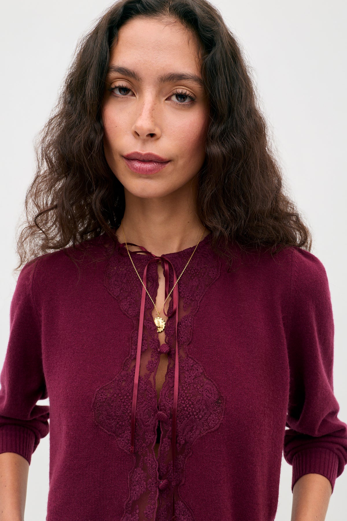 A woman with wavy brown hair wears the Tassia - Burgundy lace-trimmed sweater and a gold heart-shaped pendant necklace, standing against a plain light background.