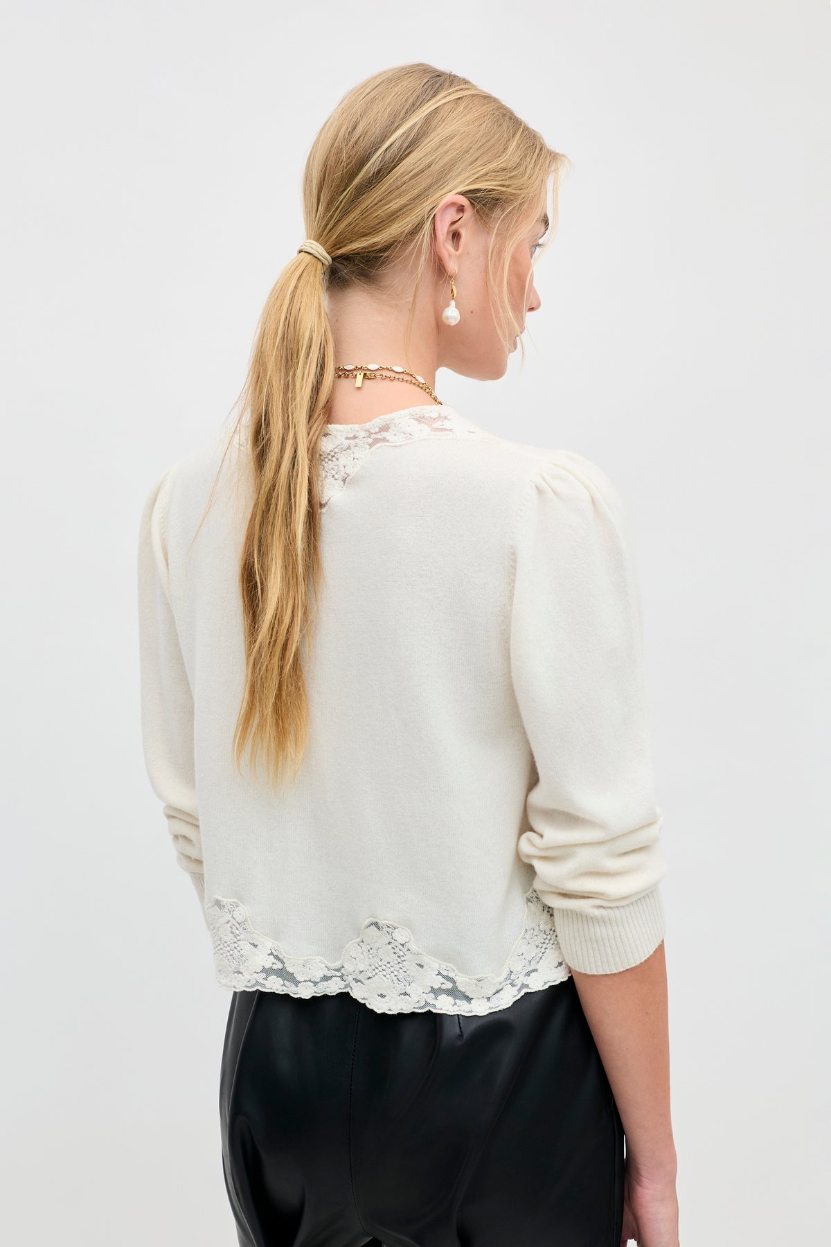 A woman with long blonde hair in a ponytail is seen from the back against a plain white background, wearing the Tassia - Cream sweater with lace trim and black pants.
