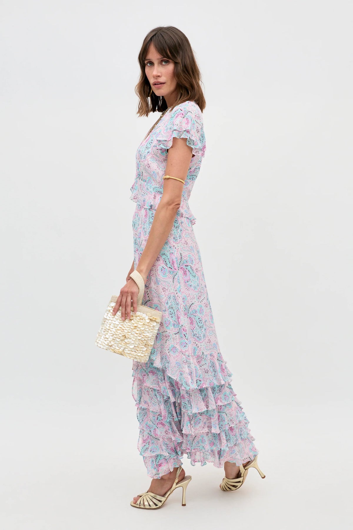 A woman stands sideways against a plain white background wearing the Thanvi - Butterfly Paisley Cream dress with ruffle details, paired with cream heeled sandals, a gold arm cuff, and a woven beige handbag.