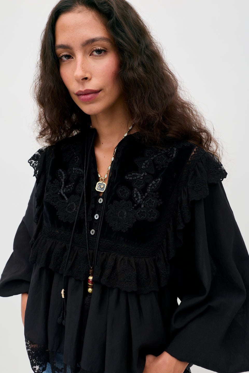 A woman with long wavy hair wears the Diann - Lilly Embroidery Black blouse, featuring delicate lace details, as she stands against a plain white background and gazes calmly at the camera.
