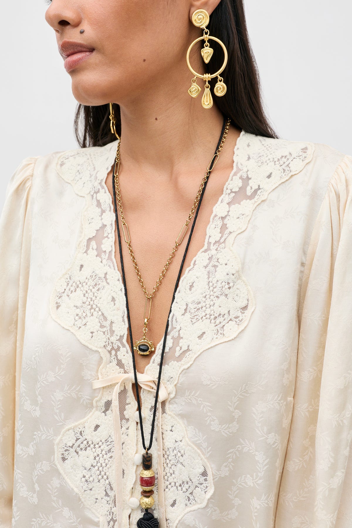 A woman wears the Tove - Floral Trail Jacquard Champagne dress, styled with layered gold necklaces and large, ornate gold earrings. The image is cropped from her shoulders to mid-chest against a neutral background.