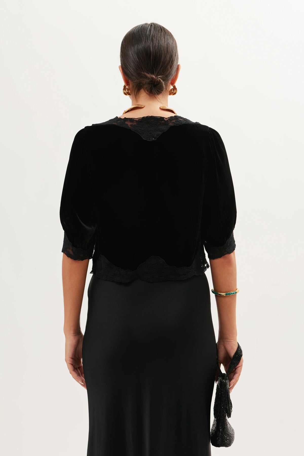 Seen from behind, a person with dark, slicked-back hair wears the Ursula - Black lace trimmed silk velvet top and a long black skirt. Gold hoop earrings, bracelets, and a small black clutch complete this vintage-inspired elegant ensemble.
