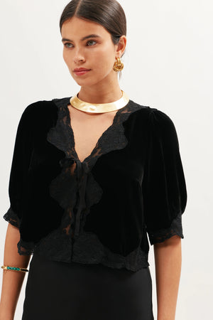 A woman with dark hair in a low bun wears the Ursula - Black silk velvet top with lace trim and ribbon tie, paired with gold jewelry and a black skirt, exuding vintage charm against a light background.