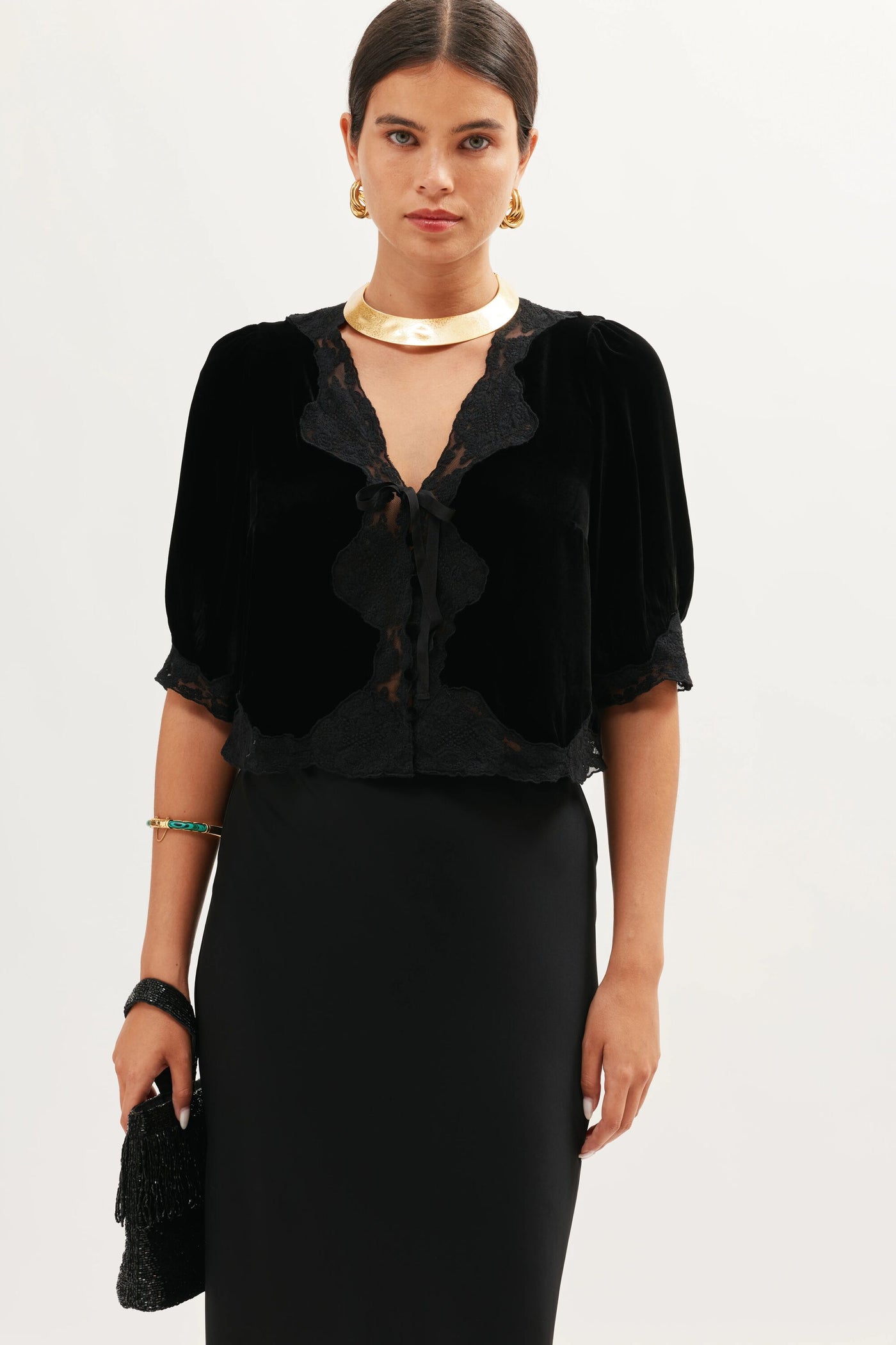 A woman with dark hair tied back models the Ursula - Black, wearing a lace-trimmed velvet blouse, black skirt, gold hoop earrings and necklace, and holding the black textured clutch against a plain white background.