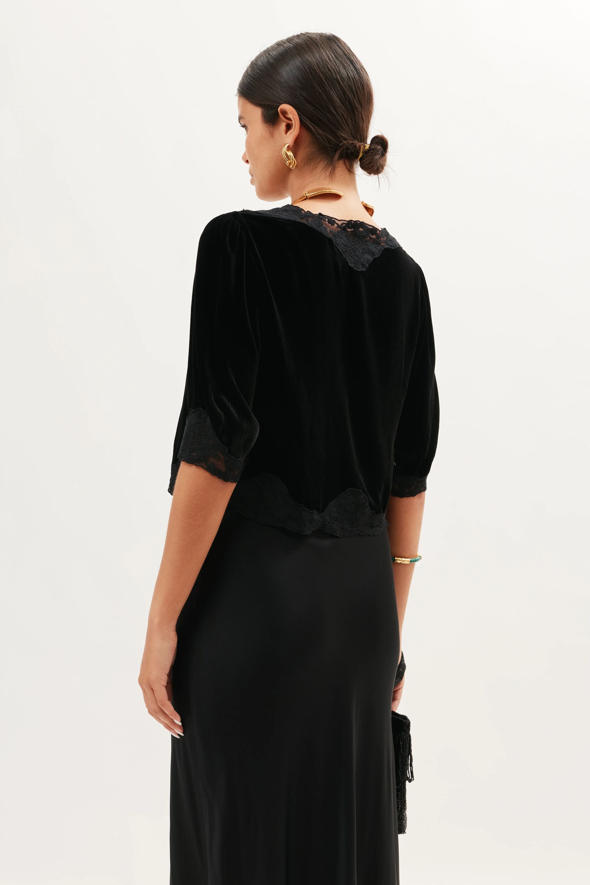 A woman faces away from the camera, wearing the Ursula - Black lace-trimmed silk velvet top with a black satin skirt. Her dark hair is in a low bun, and she holds a small black clutch for an elegant, vintage-inspired look.