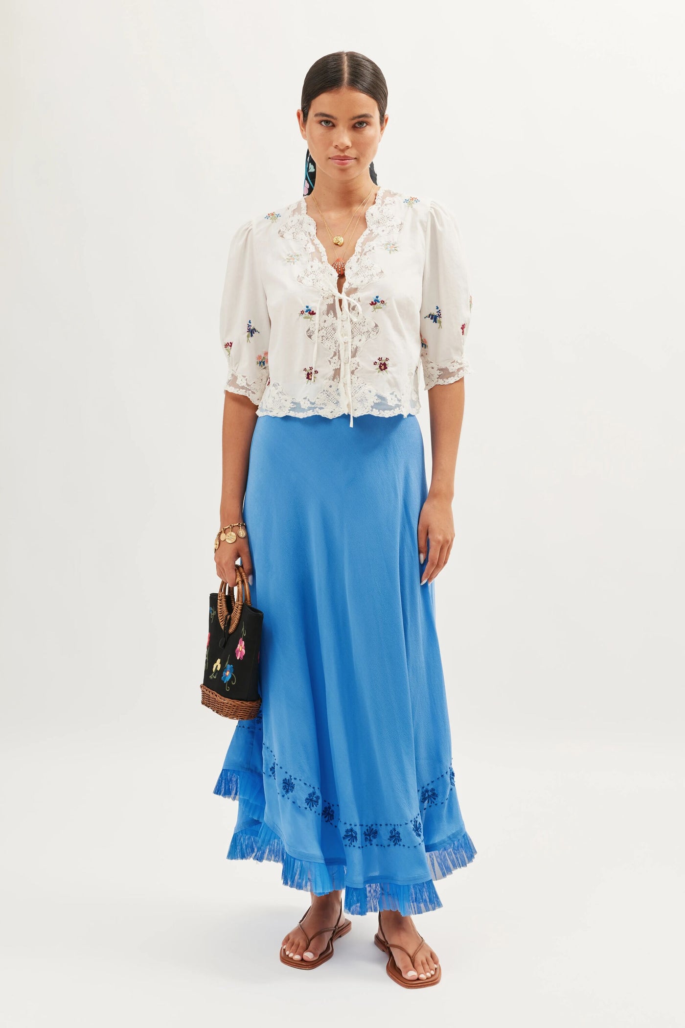 A woman stands against a plain background wearing the Ursula – Bud Embroidery White top, styled with a long blue ruffled skirt, brown sandals, and holding a black embroidered handbag for vintage charm.