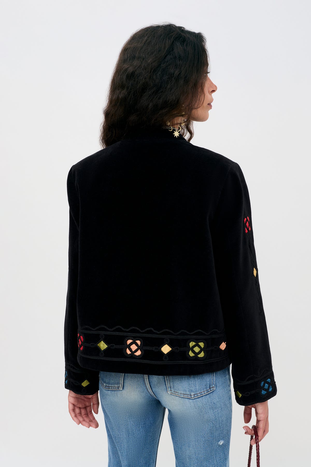 Someone with wavy dark hair, seen from behind against a plain light background, is wearing the Viktoria - Serino Applique Black jacket featuring colorful geometric embroidery along with light blue jeans.