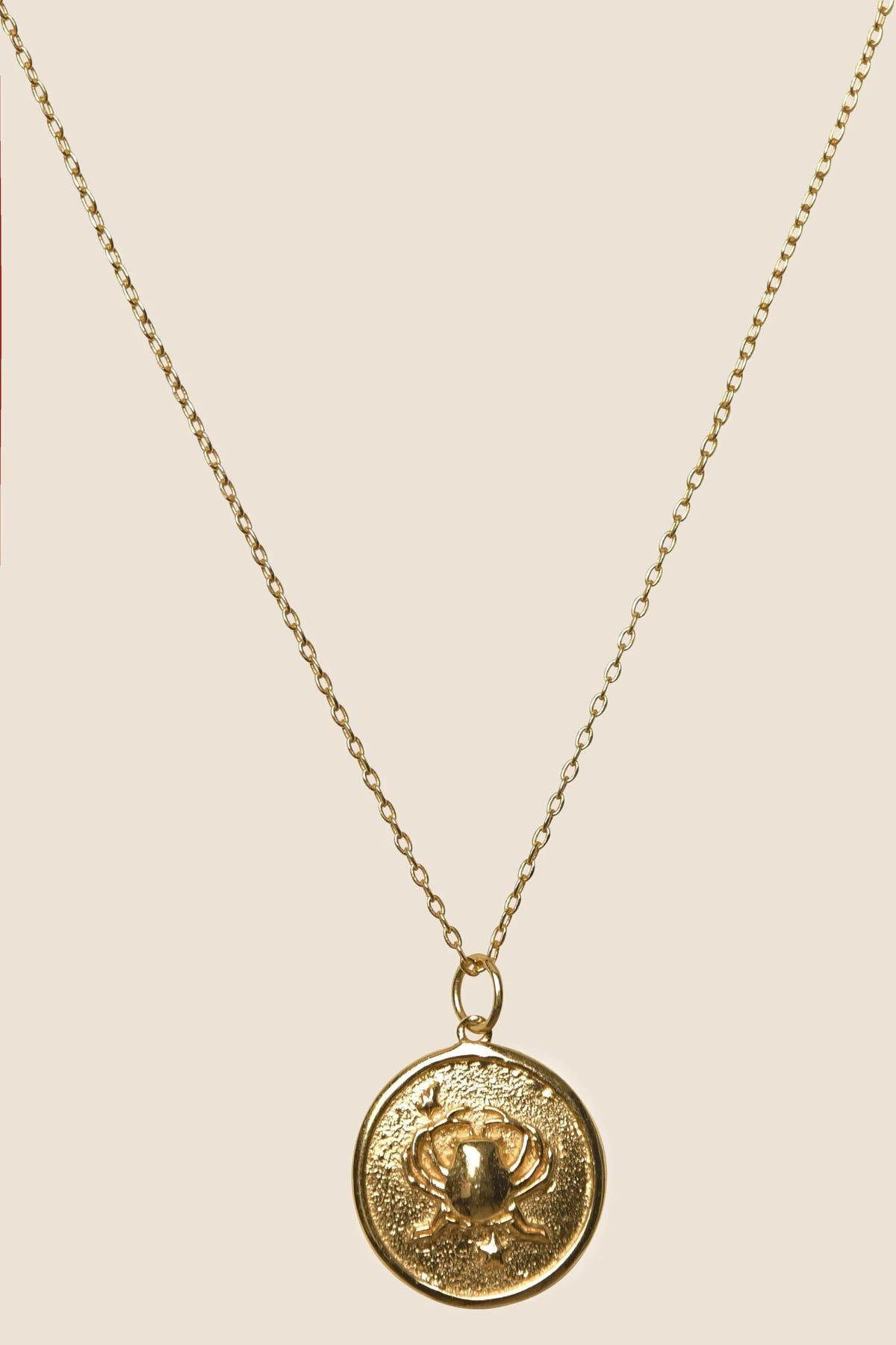 The Zodiac - Cancer necklace features gold vermeil, a delicate chain, and a round pendant engraved with a detailed crab design, shown against a simple beige background.
