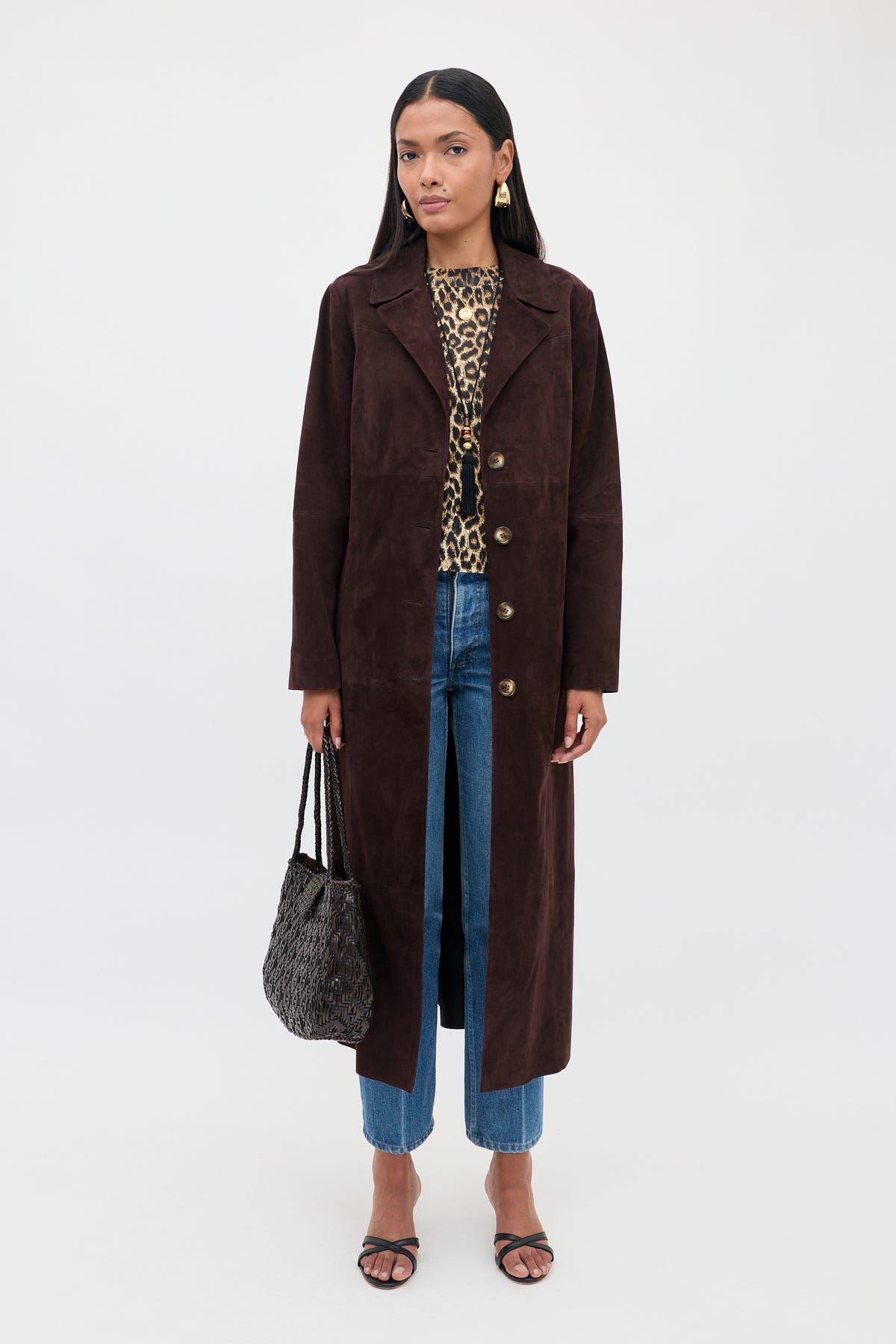 Wearing the Astraea - Chocolate suede coat over a leopard print top and blue jeans, a woman stands against a plain white background, holding a textured black bag and wearing black sandals.
