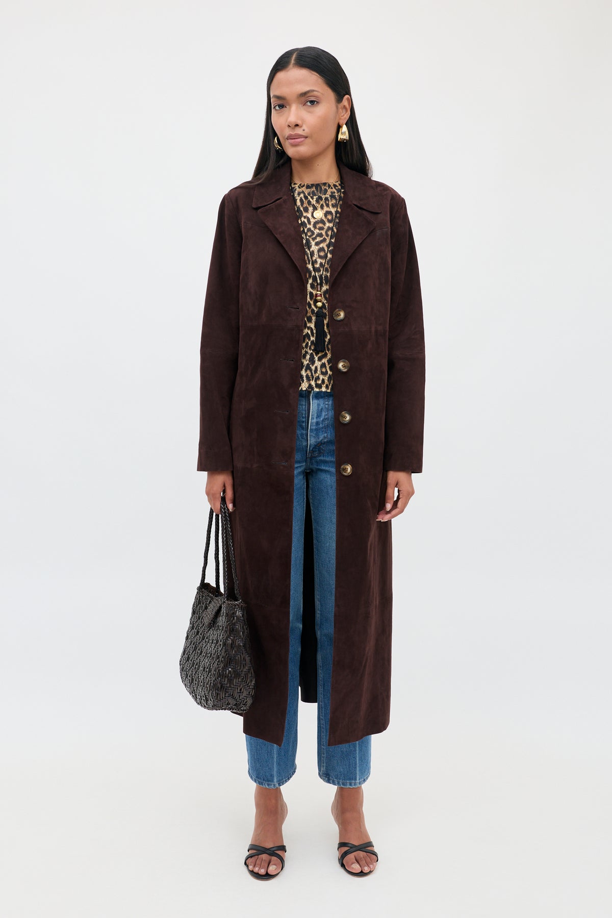 Wearing the Astraea - Chocolate suede coat over a leopard print top and blue jeans, a woman stands against a plain white background, holding a textured black bag and wearing black sandals.