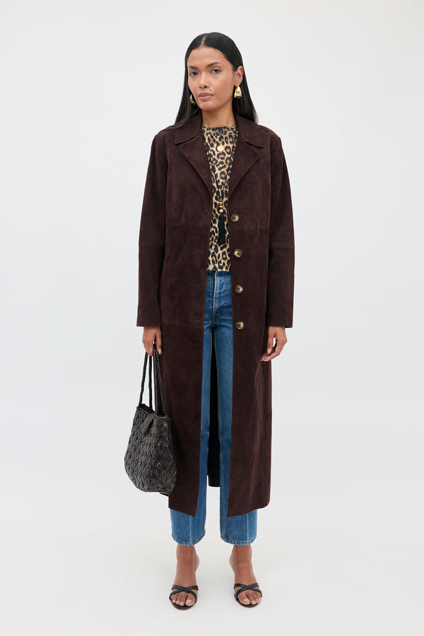 Wearing the Astraea - Chocolate suede coat over a leopard print top and blue jeans, a woman stands against a plain white background, holding a textured black bag and wearing black sandals.