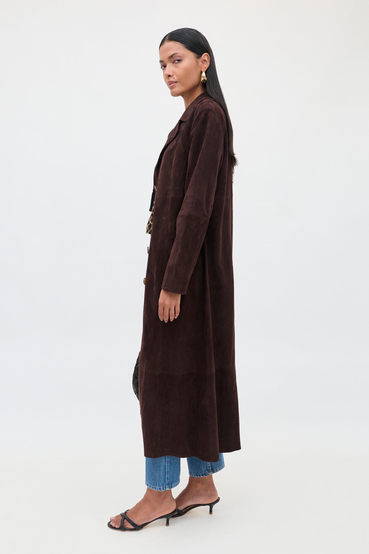 A woman wearing Astraea - Chocolate, a longline suede coat in dark brown, along with blue jeans, black heeled sandals, and gold earrings stands in profile against a plain white background.